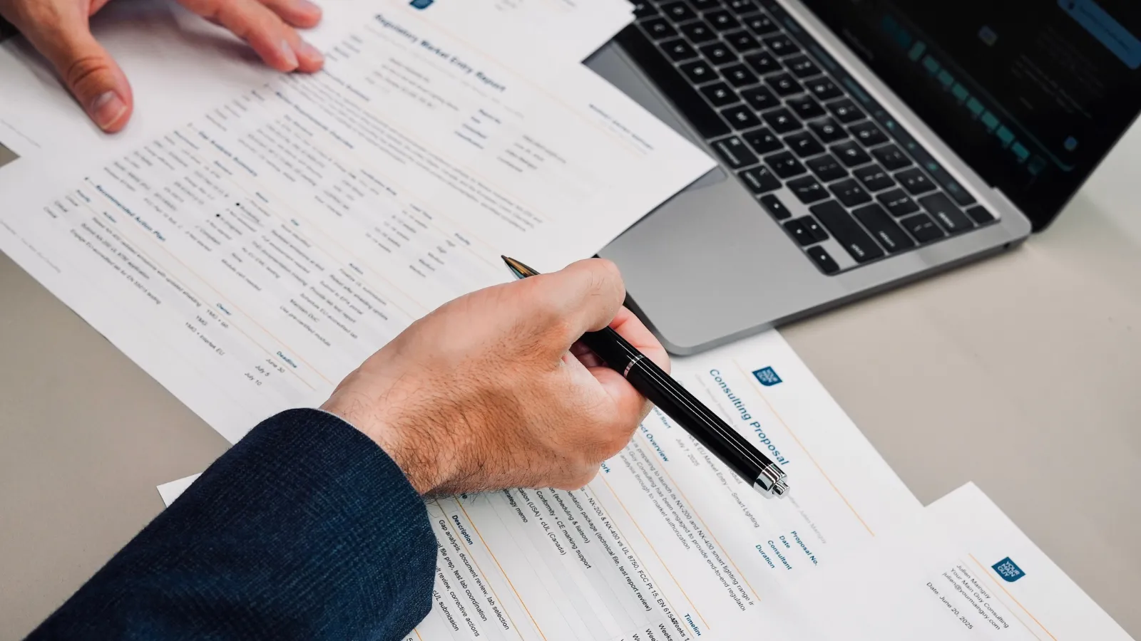 Close-up of a hand signing a regulatory consulting proposal document beside a laptop