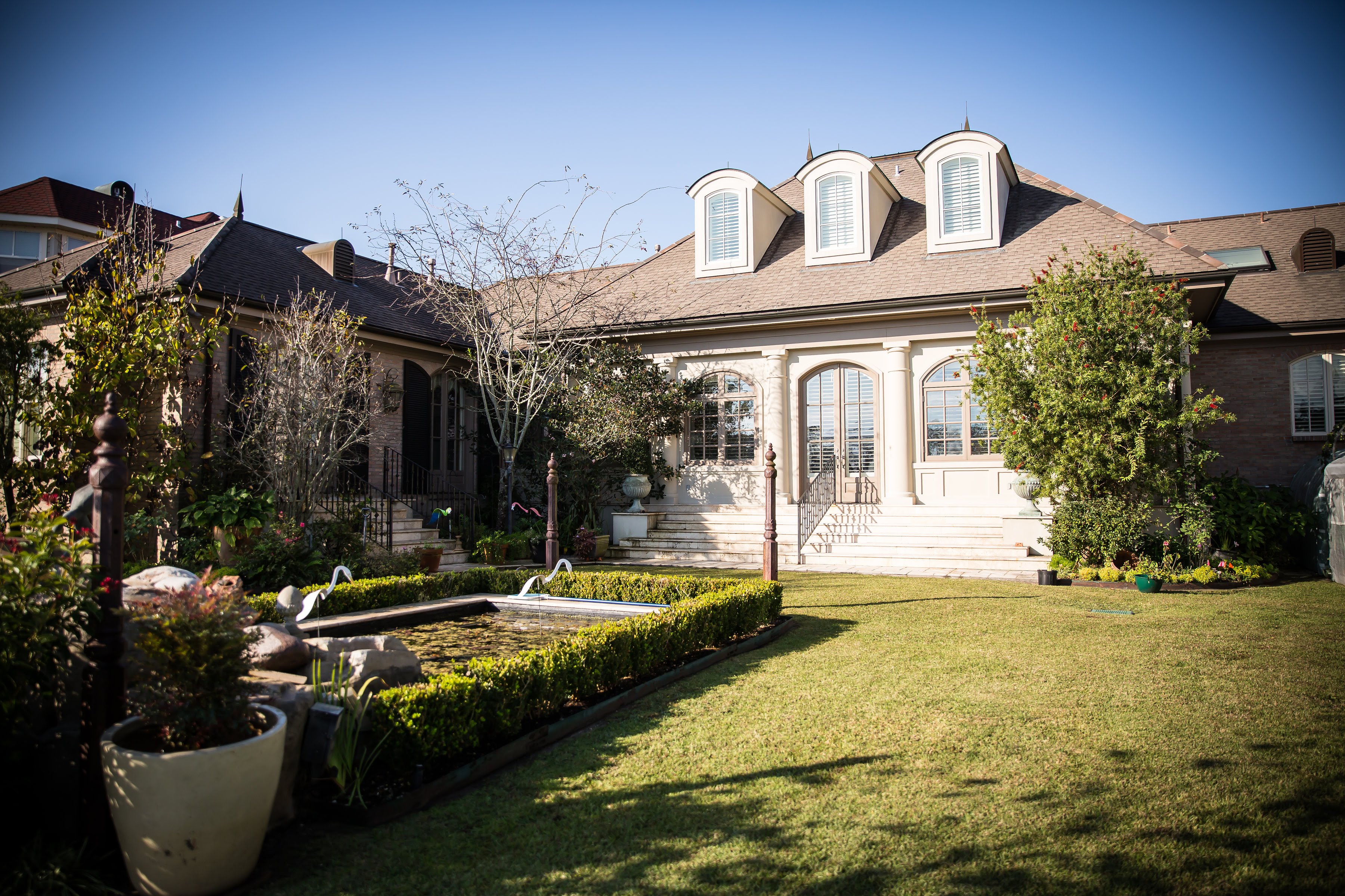 Custom French-inspired home exterior with arched dormers, formal garden, reflecting pool, and manicured lawn