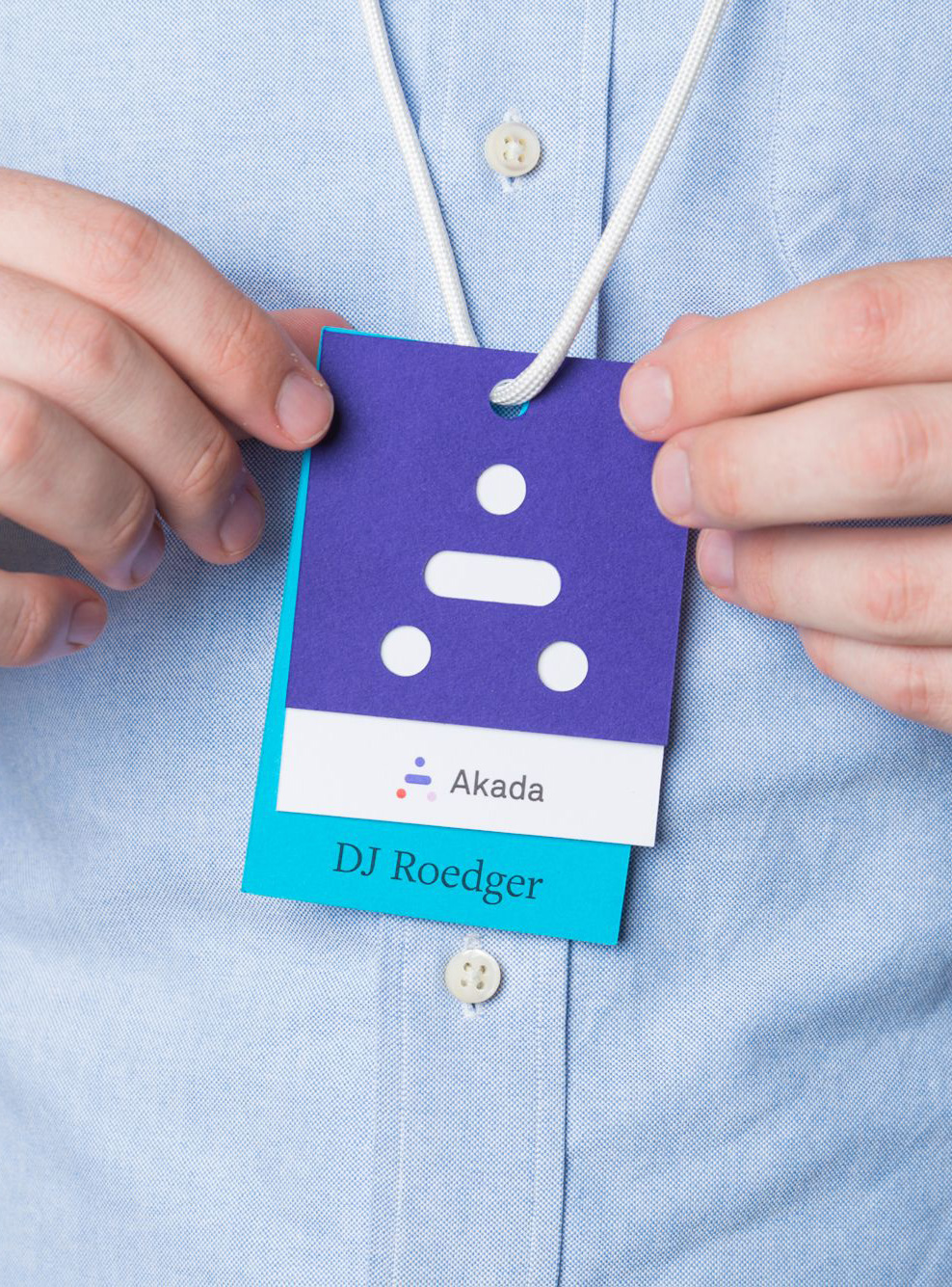 Person wearing light blue shirt holding two layered event badges, one purple with Akada logo and one blue with name DJ Roedger.
