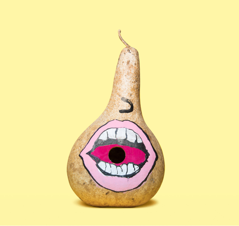 Dried gourd painted with a large open mouth featuring white teeth and a pink tongue with a hole in the center, against a pale yellow background.
