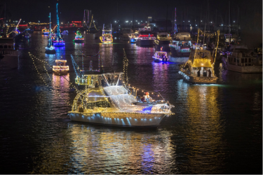 Boats decorated with colorful holiday lights parade across a harbor at night, casting bright reflections on the water.