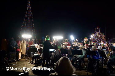 Musicians perform outdoors at night beside a Christmas tree and a tall mast decorated with colorful lights.