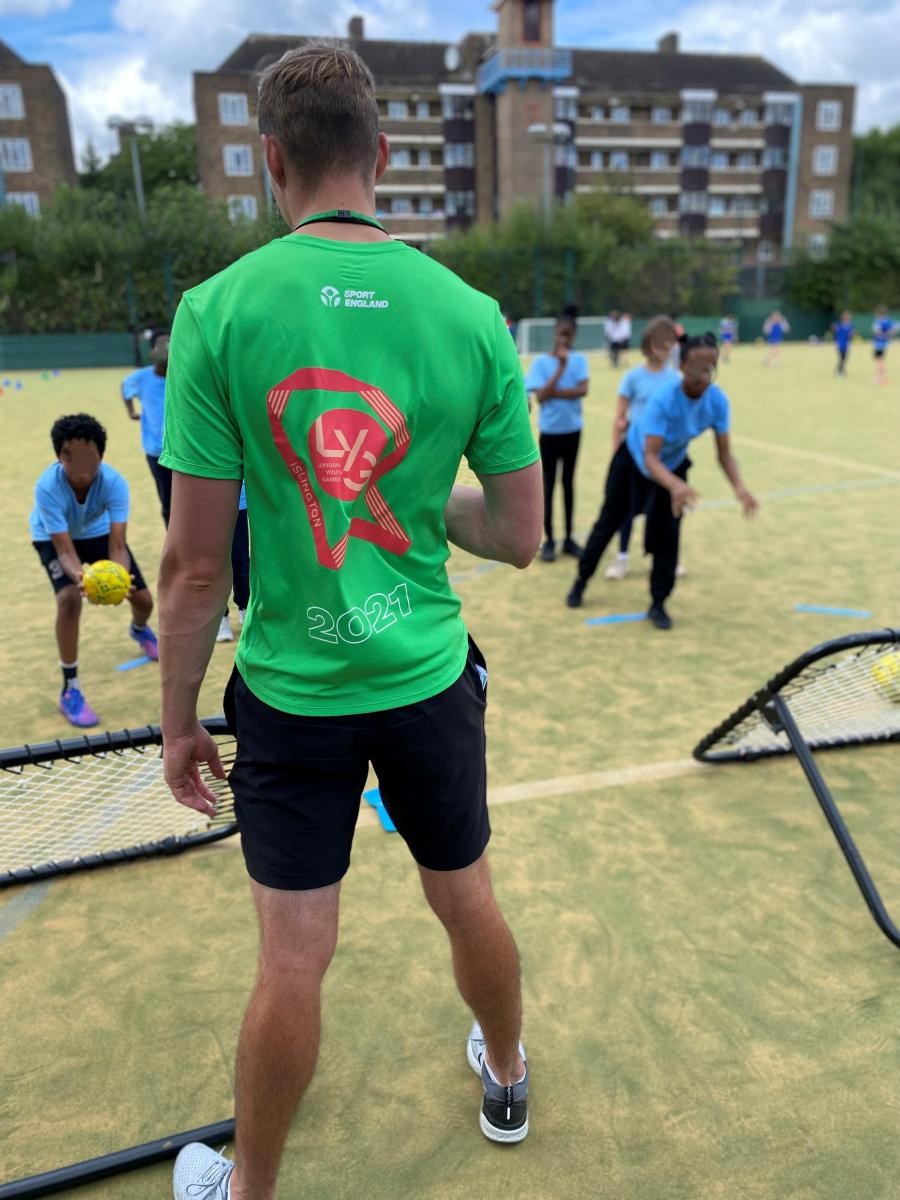 Coach in green London Youth Games shirt watching children playing Crazy Catch on outdoor pitch