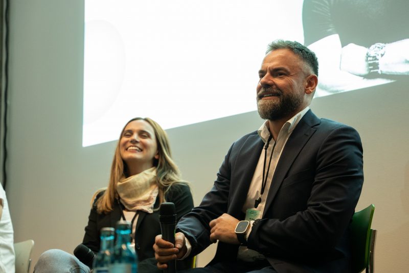 Man in a suit holding a microphone and smiling, with a woman beside him smiling as well during a panel discussion.