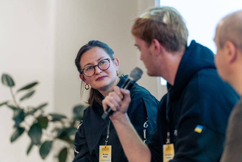 Woman with glasses listening attentively as a man speaks into a microphone during a panel discussion.