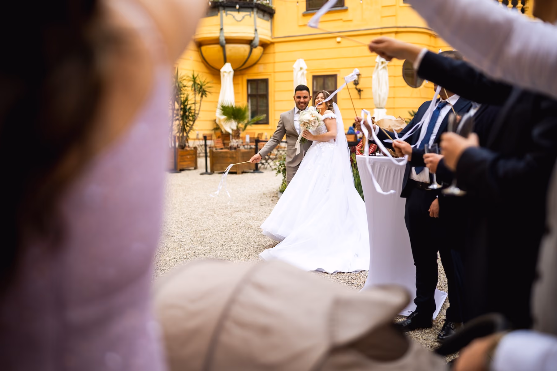 Novomanželia s kyticou medzi hosťami mávajúcimi stuhami na nádvorí.