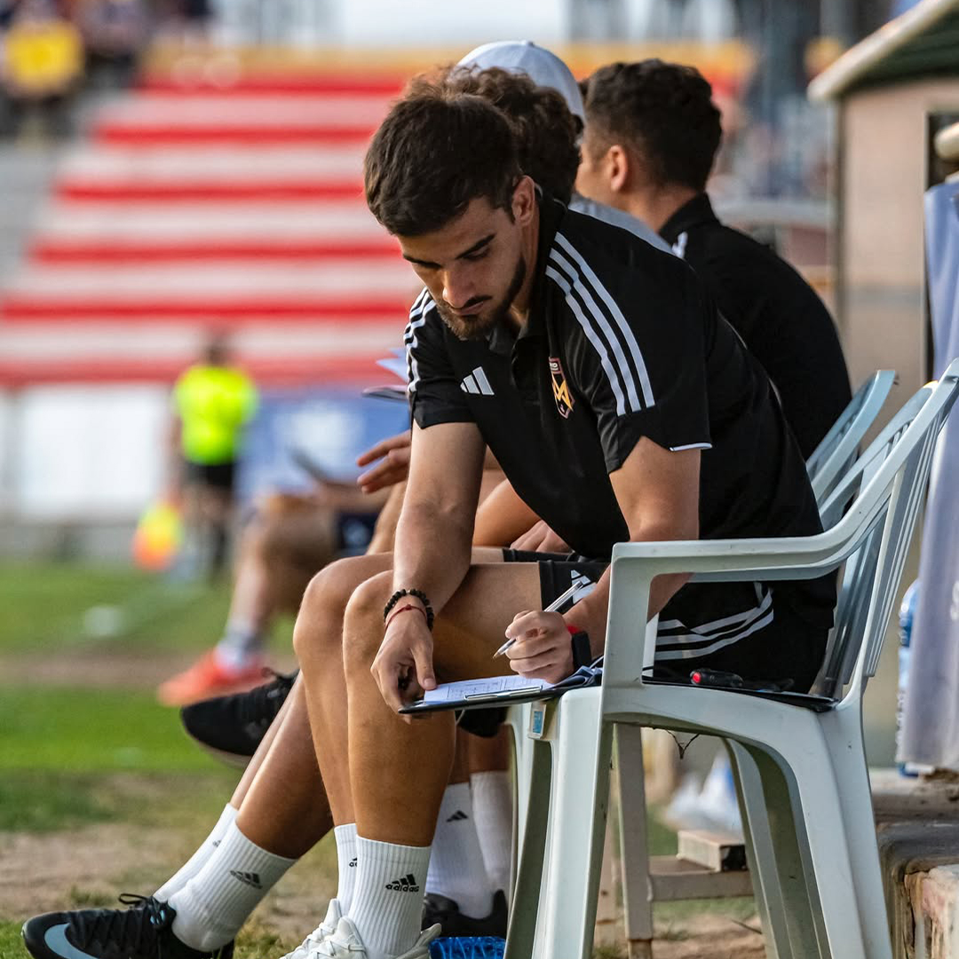 A Spanish football academy coach taking notes during a soccer game.