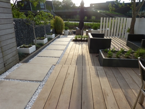 Een hardhouten vlonder met een pad van keramische tegels in wit grind, drie plantenbakken op rij en verticale witte palen.