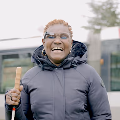 Front view of a person holding a white cane and wearing Envision Glasses outside.