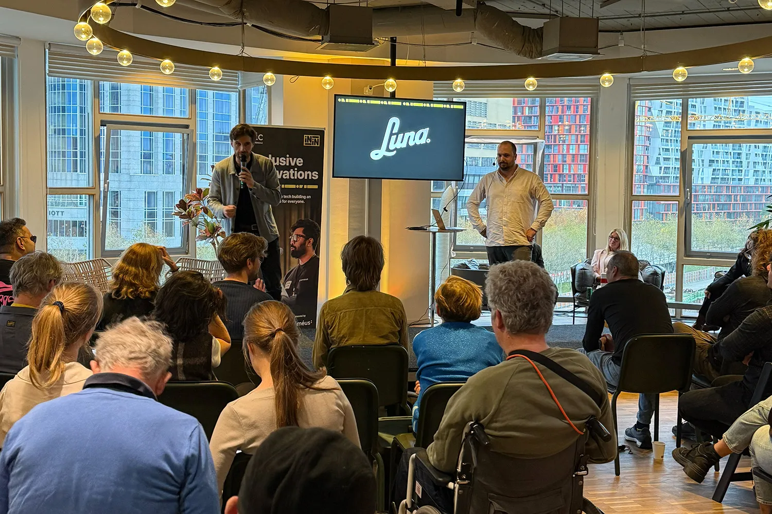 A man stands on stage holding a microphone and speaking to an audience while another man stands nearby with hands on hips. Behind them, a screen displays the word “Luna” in cursive script. The event appears to be part of the Inclusive Innovations initiative, as indicated by a vertical banner on the left. The audience is seated facing the speakers, with one person in a wheelchair visible in the foreground. Large windows in the background reveal modern city buildings and colorful high-rises. The setting is well-lit and professional.