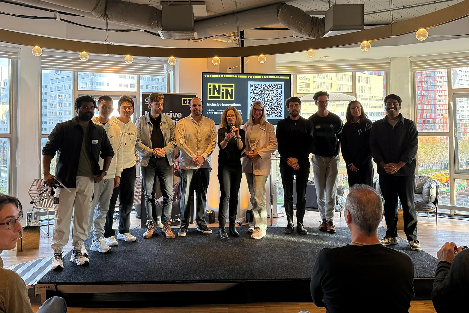 A group of thirteen innovators stand on a small stage in front of large windows at an indoor event. They are posing for a photo. Behind them is a screen displaying the logo of Inclusive Innovations and a large QR code. Two banners flank the screen, and city buildings are visible through the windows. Audience members are seated in the foreground.