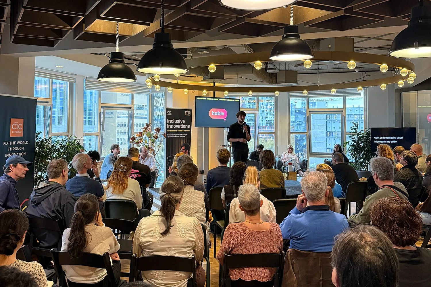 A man stands on stage holding a microphone and presenting to a seated audience at an indoor event. Behind him, a screen displays the logo “hable” in a red speech bubble. Banners for "Inclusive Innovations" and "CIC Rotterdam" are visible on the left side of the room. Additional people are seated on stage, and a second screen on the right displays text about translating visual information into audio. Large windows in the background reveal city buildings. The event has a modern, inclusive, and tech-focused atmosphere.