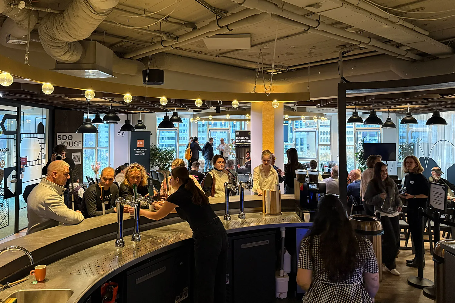 People gather around a curved bar counter in a modern indoor space during a networking or break session at an event. A woman behind the bar serves drinks while attendees socialize, chat, and wait in line. In the background, banners for CIC Rotterdam and Inclusive Innovations are visible near large windows with city buildings outside. Hanging lights and exposed ceiling pipes contribute to the industrial, creative atmosphere. The setting appears informal yet professional, with a focus on innovation and collaboration.