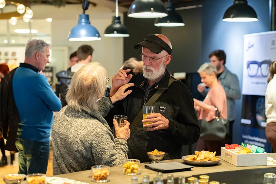 A group of people socializing at the Inclusive Innovations 2025 event. In the foreground, an older man and woman engage in conversation while holding drinks, standing near a table with bowls of snacks and chips. The background features other guests mingling, with a display and promotional poster for smart glasses visible on the right. The setting is well-lit with hanging industrial-style lights.