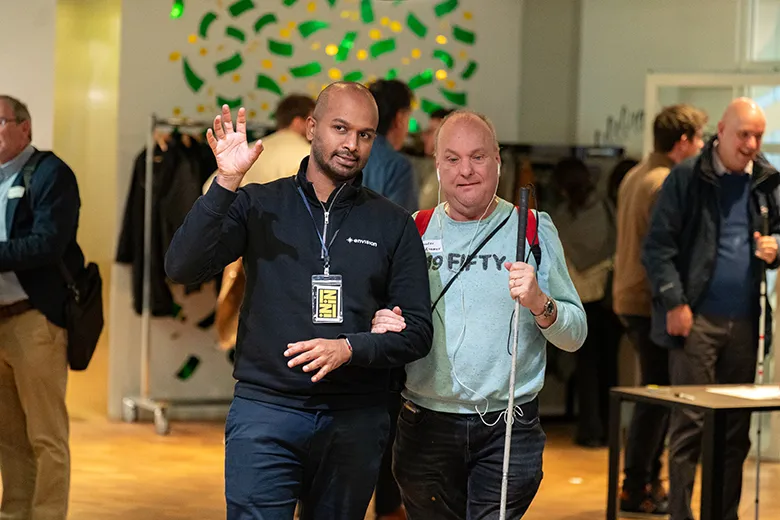 A visually impaired man holding a white cane is being guided through a busy indoor event space by a man wearing a name badge and moving his hands, hinting at an ongoing conversation. The guide has his arm linked with the man he is assisting. Several other people are visible in the background, some also holding white canes. The environment is well-lit with a modern design and a wall decorated with green and yellow shapes.