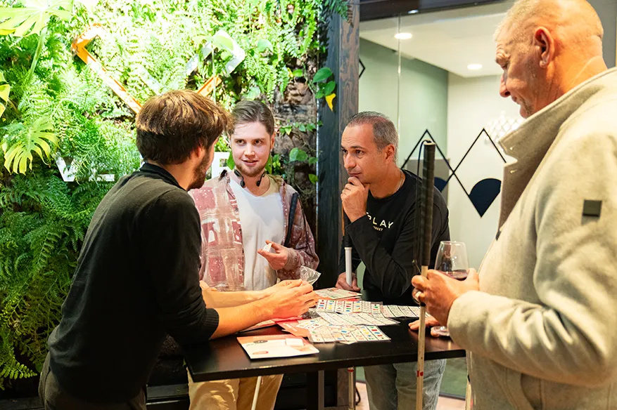 Four men are gathered around a high table engaged in a conversation. Three of the men are holding long black-and-white canes, indicating visual impairment. One of them also holds a wine glass. The setting is a brightly lit indoor space with a lush green vertical plant wall in the background.