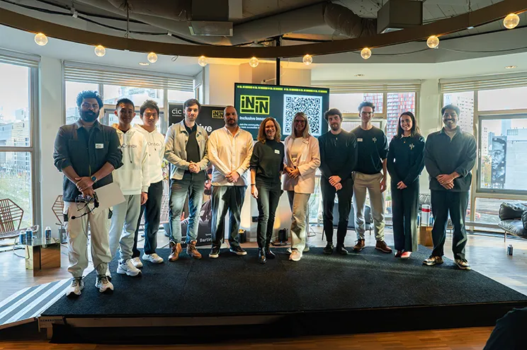 A group of thirteen innovators stand on a small stage in front of large windows at an indoor event. They are posing for a photo. Behind them is a screen displaying the logo of Inclusive Innovations and a large QR code. Two banners flank the screen, and city buildings are visible through the windows. Audience members are seated in the foreground.
