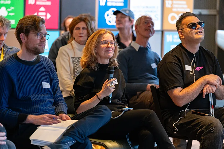 A group of people seated and attentively listening during the Inclusive Innovations 2025 presentations. A woman in the center holds a microphone and smiles, while a man on her left takes notes in a notebook. On her right, a man wearing sunglasses and earphones appears engaged. Behind them, other attendees are visible, and colorful banners displaying icons and text are mounted on the wall.