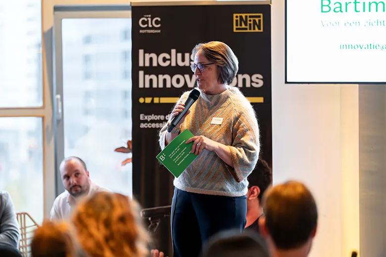 A woman stands speaking into a microphone at an indoor event, holding a green booklet in her other hand. She is positioned in front of a banner that reads “Inclusive Innovations” and “Explore our accessible future.” Audience members are seated in the foreground, and a presentation screen displaying the word “Bartiméus Fonds” is partially visible to the right. The setting appears to be a professional event focused on accessibility or inclusion.
