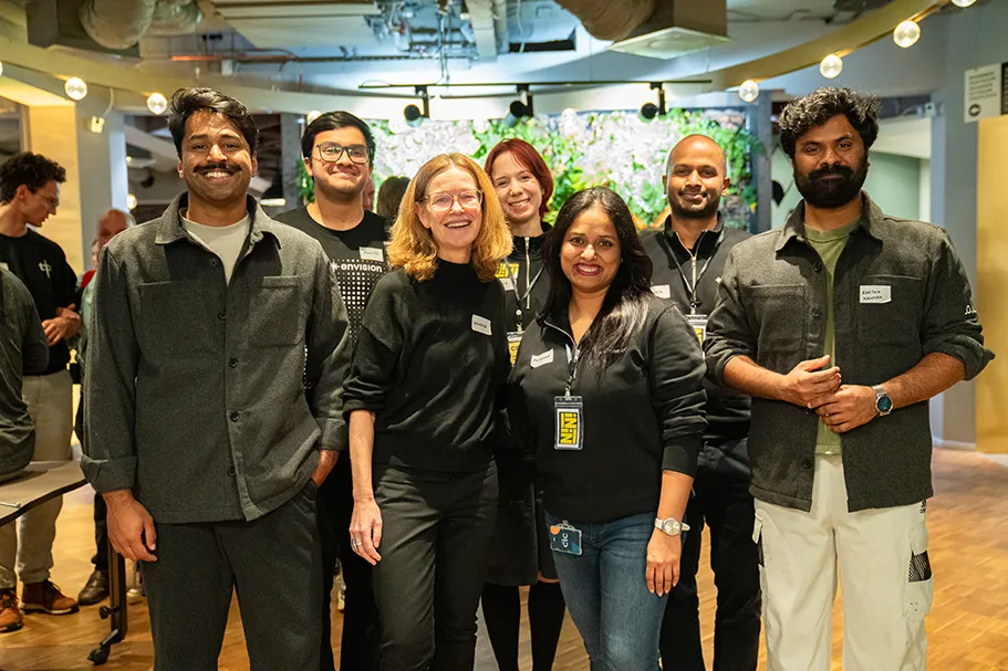 A diverse group of seven smiling people stands together indoors, posing for a photo at a professional or networking event. They are dressed in casual to business-casual attire and wearing name badges. The setting is well-lit, with a modern interior design and a digital green wall display in the background. Other attendees are visible in the blurred background.