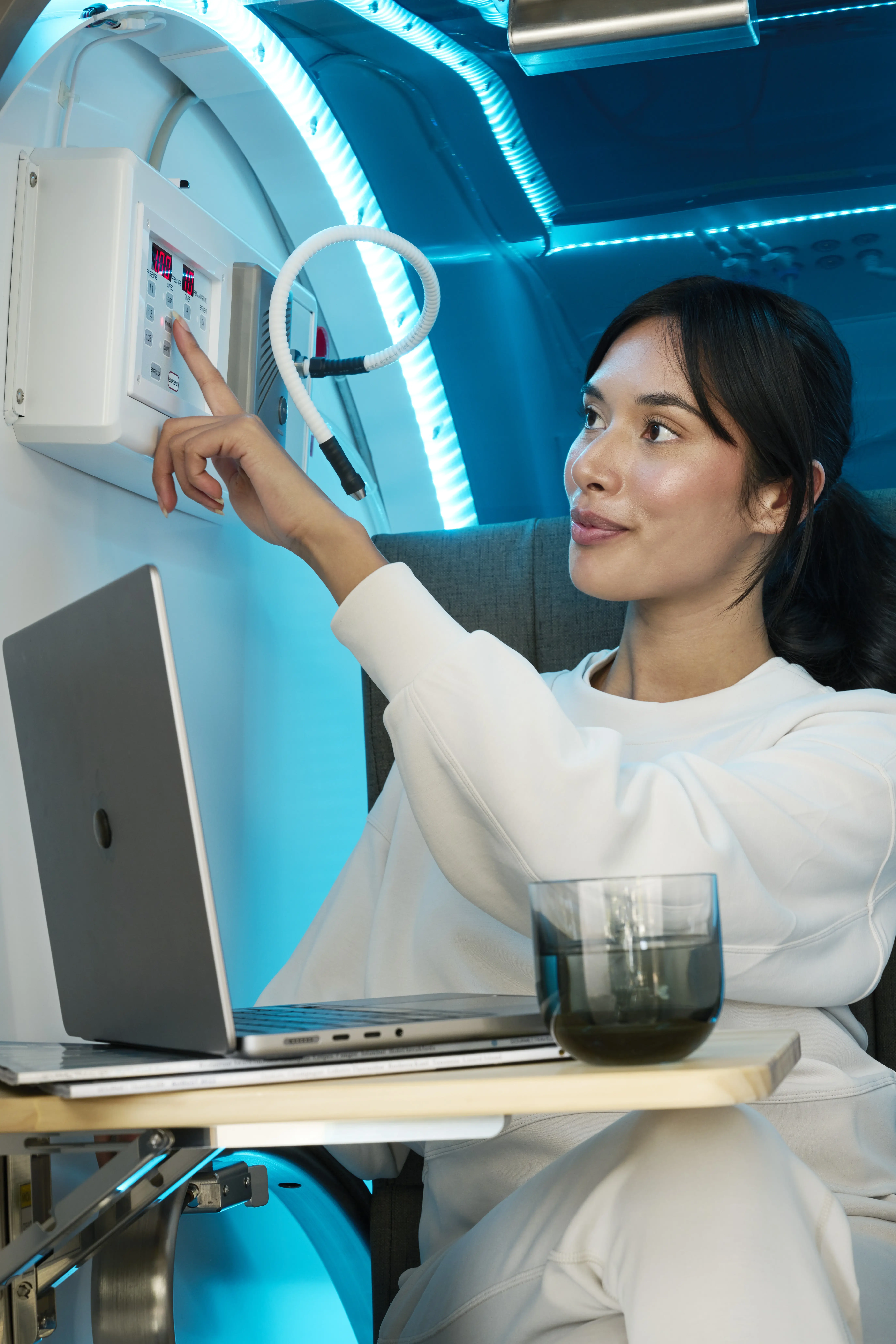 Woman in a white sweatshirt adjusting controls on a hyperbaric oxygen therapy chamber with a laptop and a glass of water on a tray table.