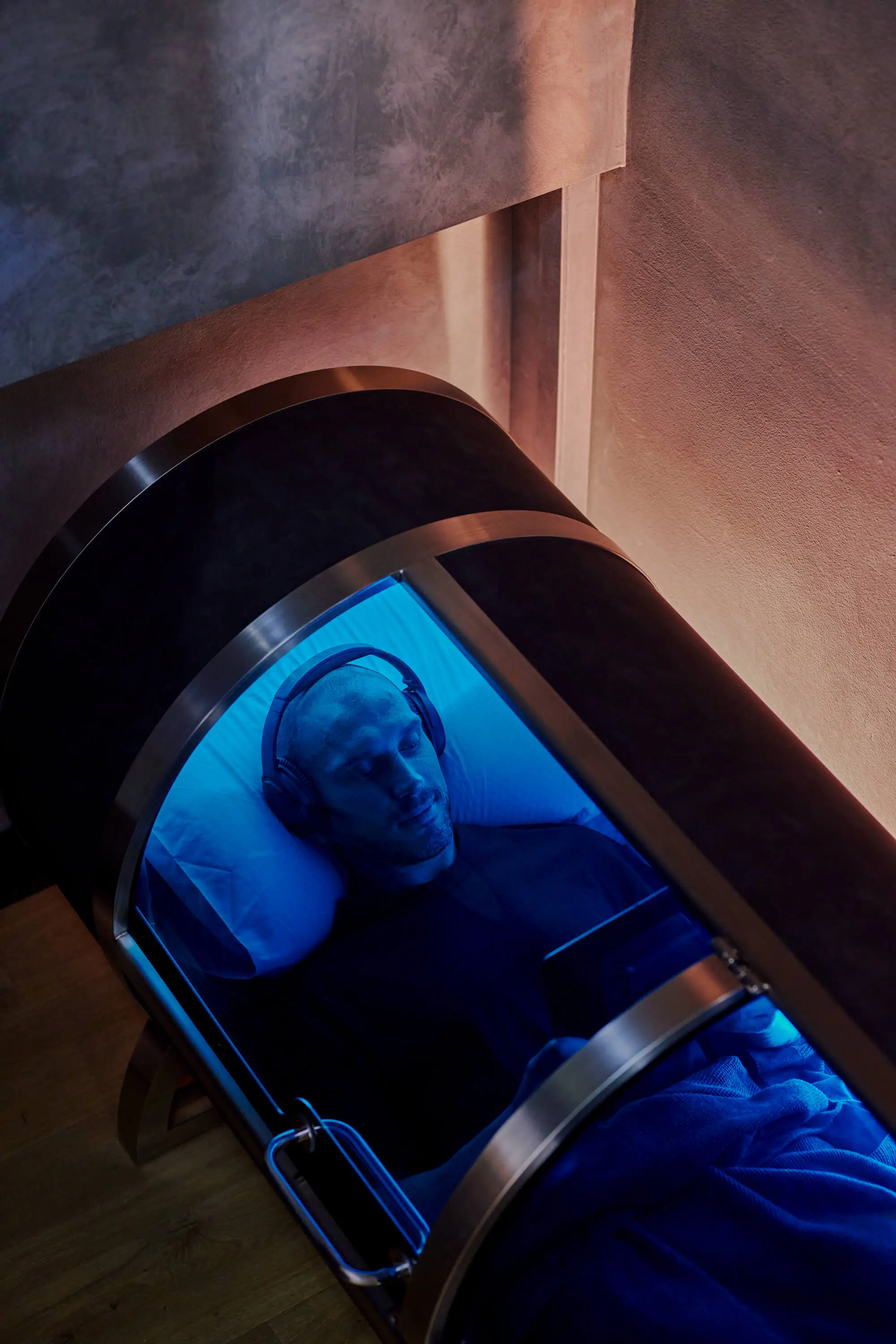 Man wearing headphones lying inside a hyperbaric oxygen therapy chamber with blue lighting, holding a tablet.