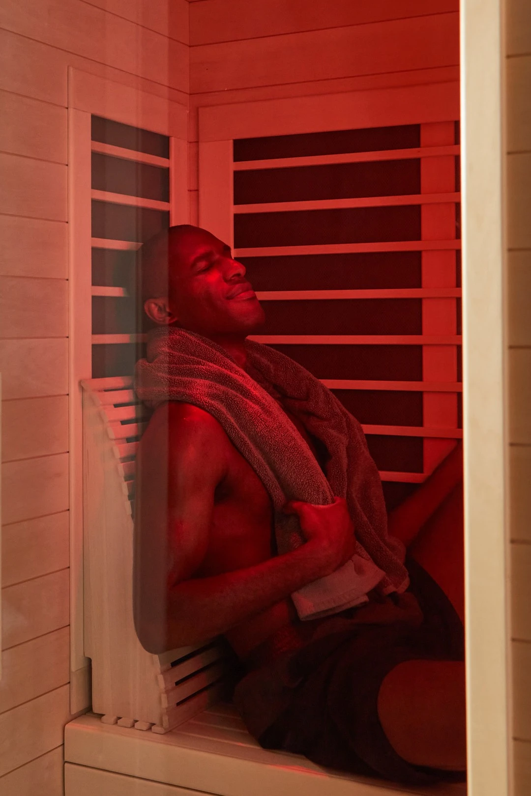 A man relaxing inside a sauna with a towel around his neck and eyes closed, bathed in red light.