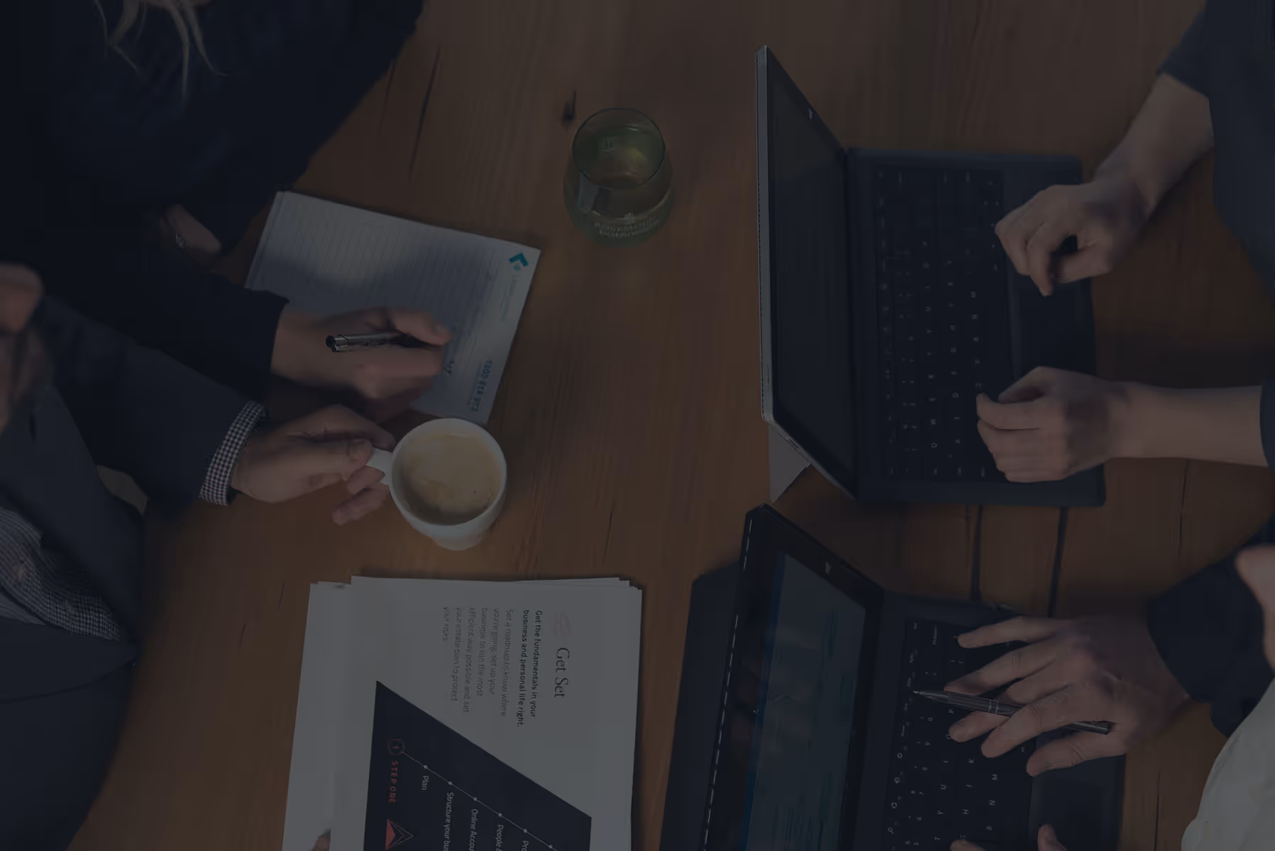 Overhead view of a team collaborating around a wooden table with laptops, printed documents, handwritten notes, and coffee during a business meeting.