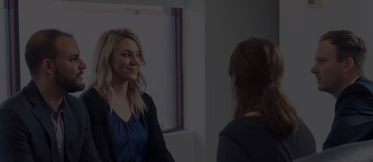 Four professionals engaged in a discussion in an office setting, with two facing forward and two with backs to the camera.