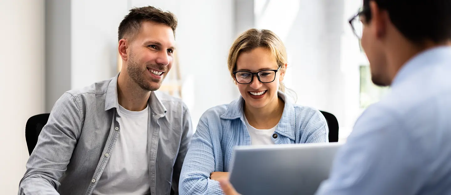 Smiling man and woman looking at a laptop screen held by another person in an office setting.