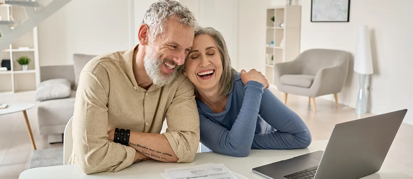 Smiling mature couple leaning on a table with paperwork and laptop, sharing a joyful moment in a cozy living room.