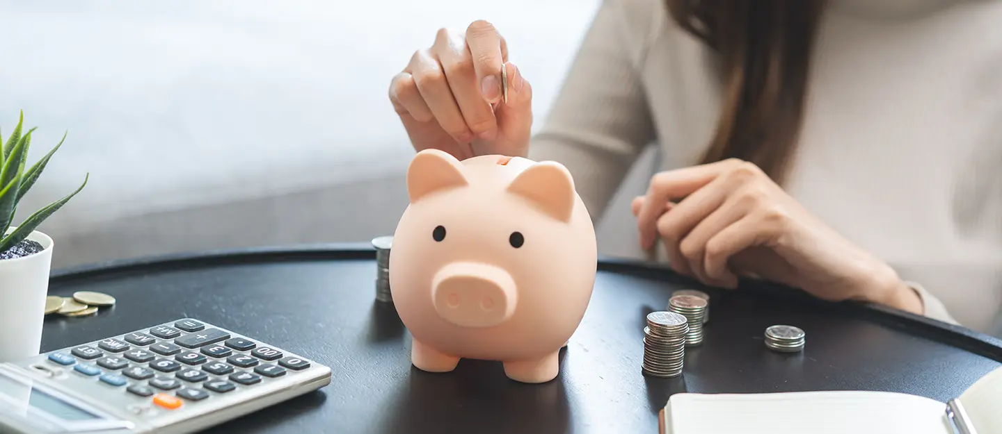Person placing a coin into a pink piggy bank on a black table with a calculator, stacks of coins, a plant, and an open notebook.