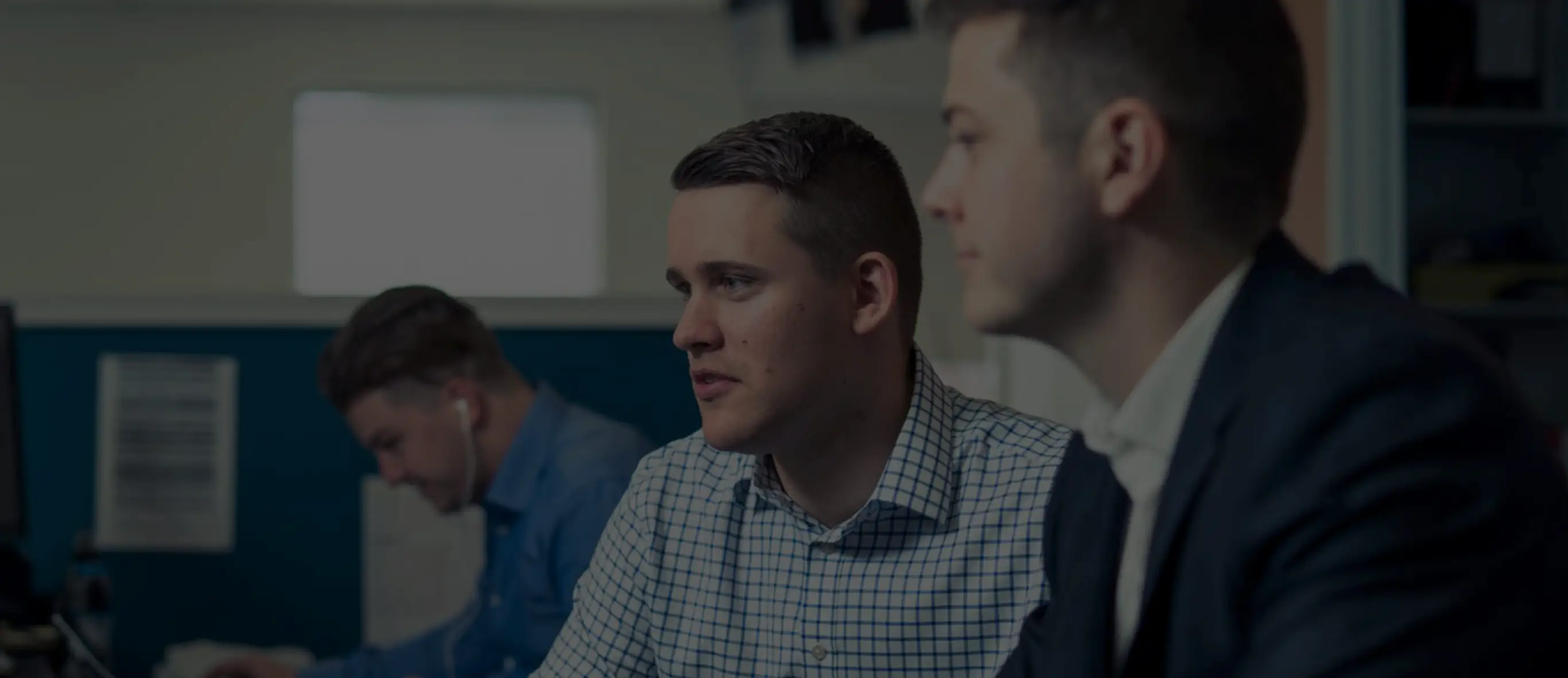 Three men focused on work in an office, with one wearing a checkered shirt and another in a suit.