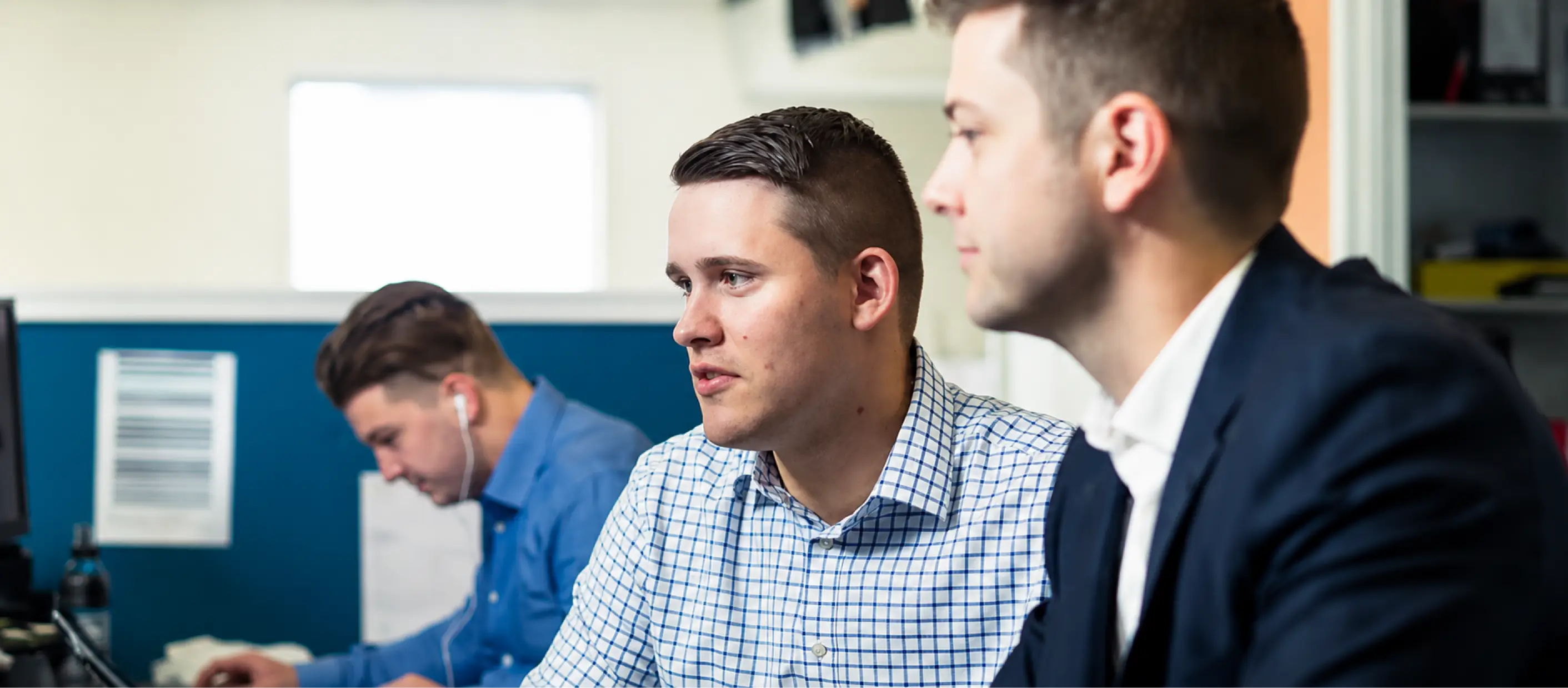 Three men working together in an office meeting space, focused on computer-based tasks and discussion.