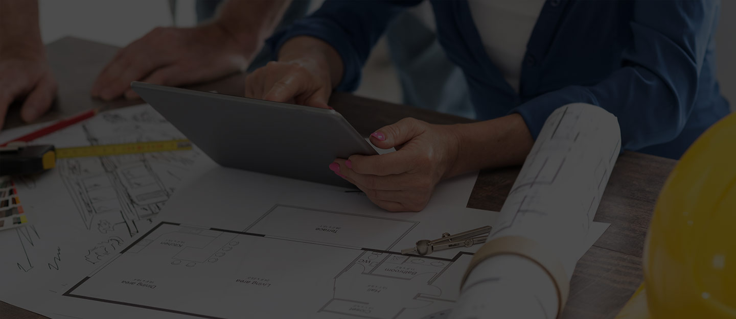 Person holding tablet over architectural blueprints with a rolled plan and yellow hard hat on table.