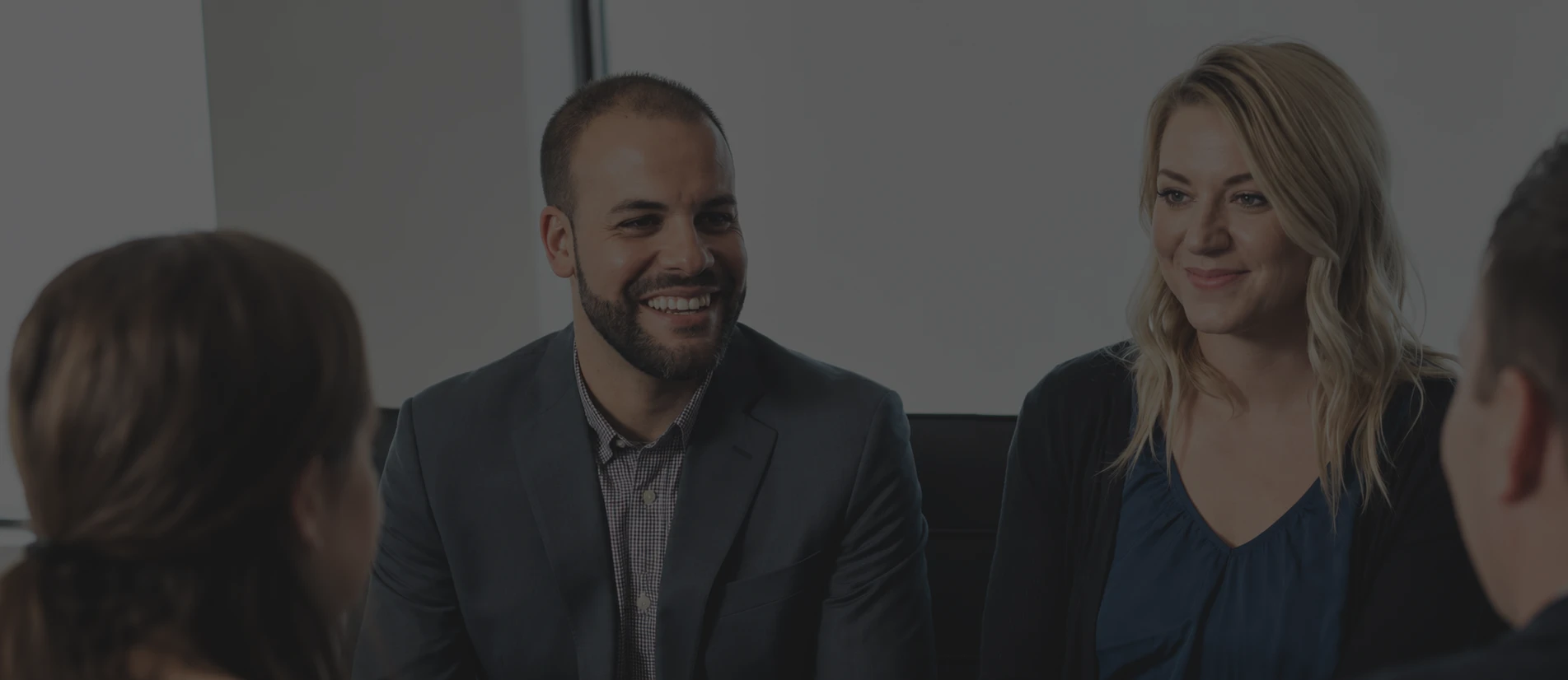 Two professionals smiling during a business meeting, seated across from colleagues in a bright office setting.