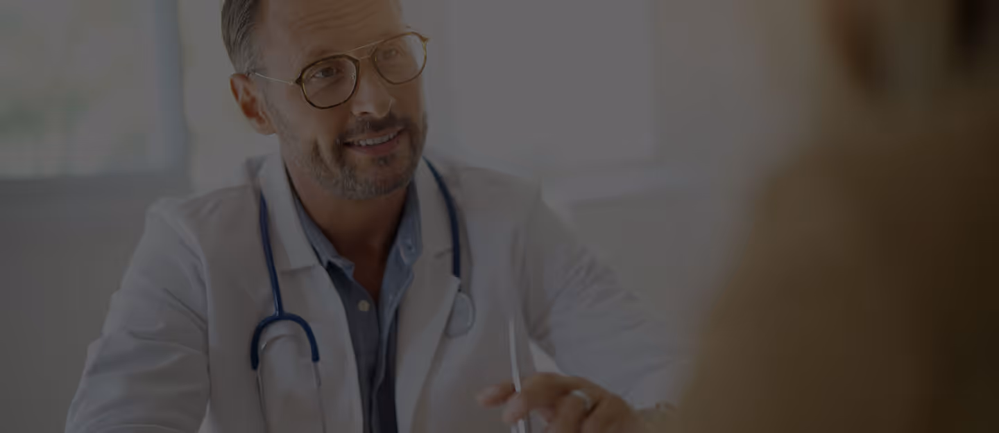A doctor standing with a chart or tablet while speaking with someone off-camera.