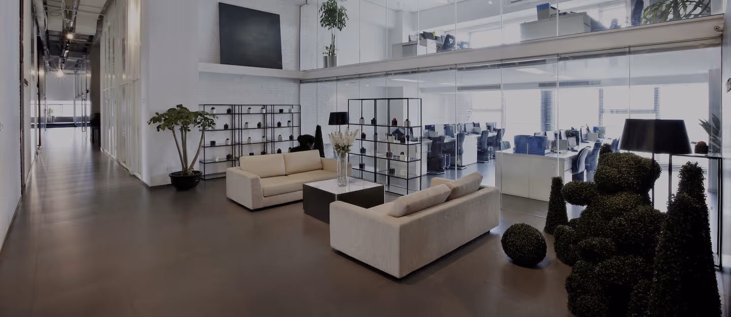 Modern office hallway with sofas, greenery, and a glass partition leading to an open plan office