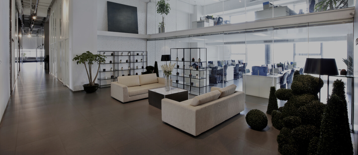 Modern office hallway with sofas, greenery, and a glass partition leading to an open plan office