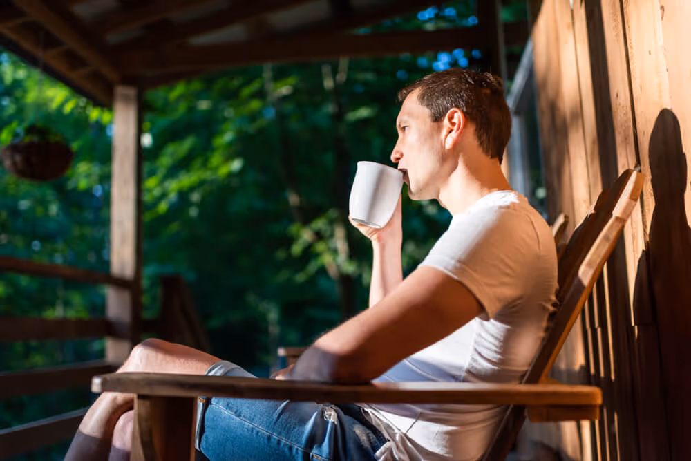 A man relaxes on the porch of his home in the early morning sun, drinking a cup of coffee.