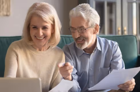 An older couple sitting on a couch, looking at a laptop and holding papers, smiling and pointing at the screen.