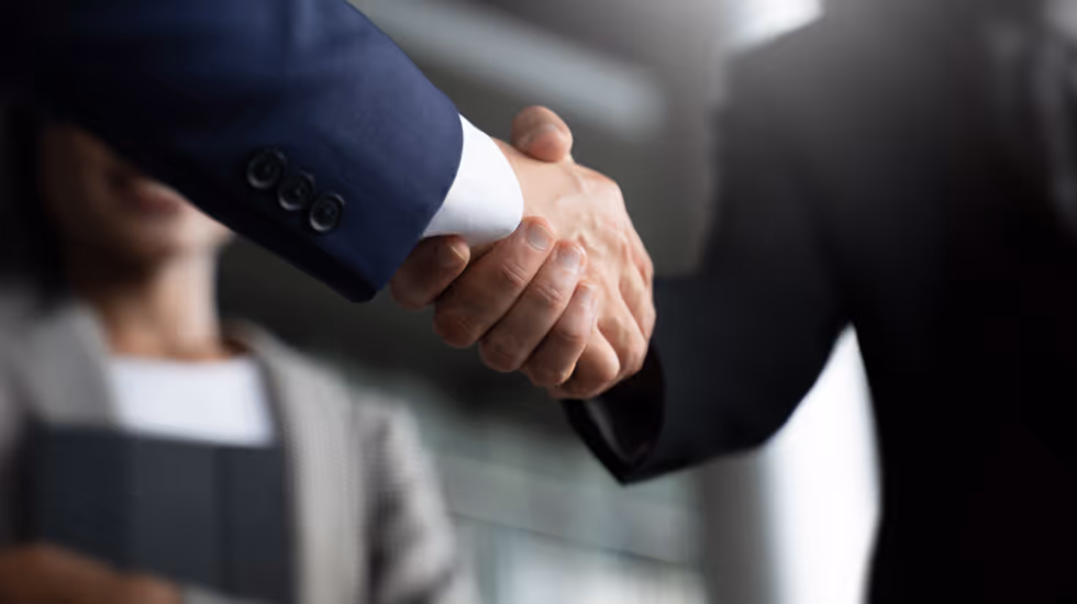 Two professionals in business attire shake hands, symbolising a formal agreement or partnership, with a third person smiling in the background.