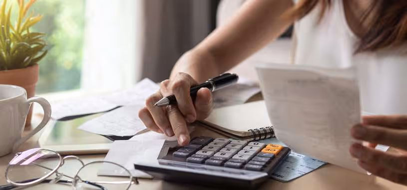 A closeup of a woman’s hands as she uses a calculator, pen and paper to work through her finances.