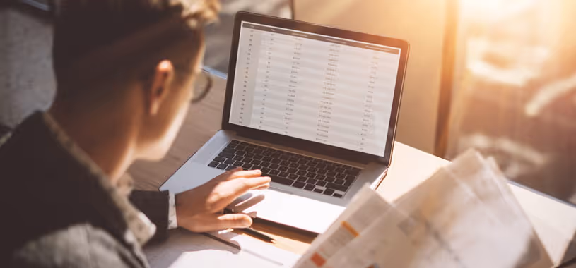An over-the-shoulder shot of a financial advisor looking over data on his laptop, as well as on printed sheets before him.