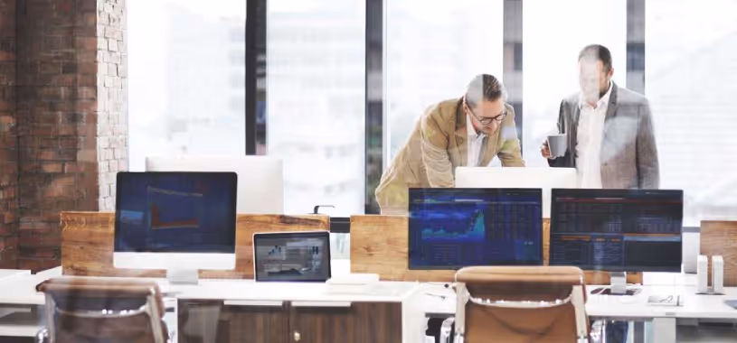 Two financial advisors in an office, looking at a computer screen showing the data of a client they’re working for.