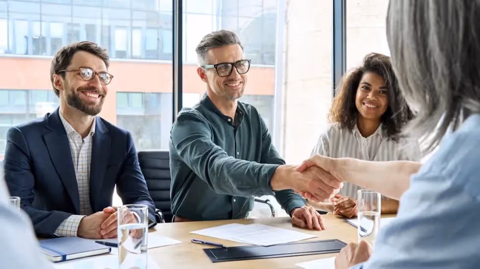 Group of business professionals smiling and shaking hands during a meeting in a bright office with large windows.