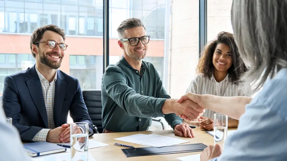Group of business professionals smiling and shaking hands during a meeting in a bright office with large windows.