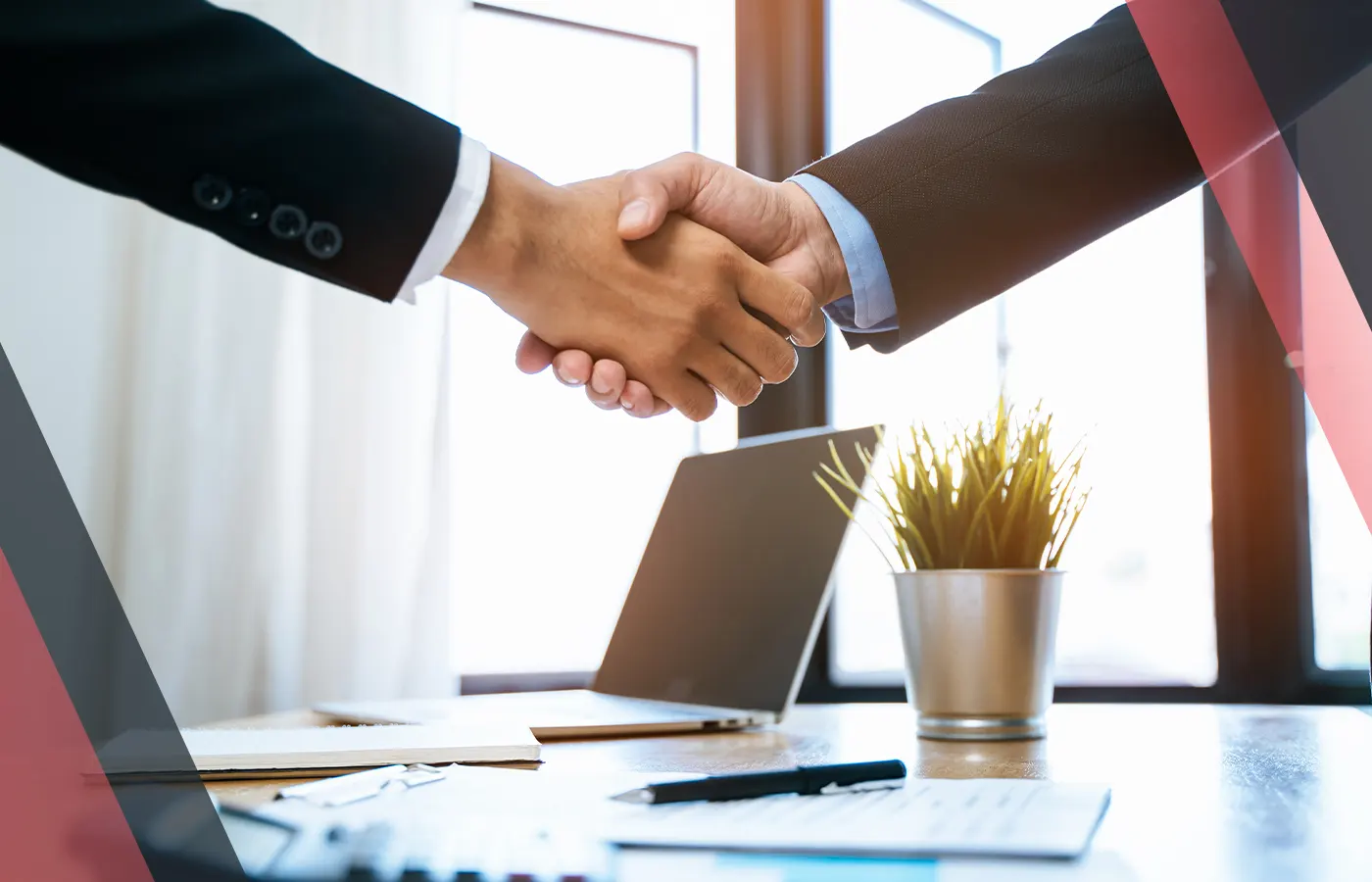Two businesspeople shaking hands across a desk with a laptop and documents.