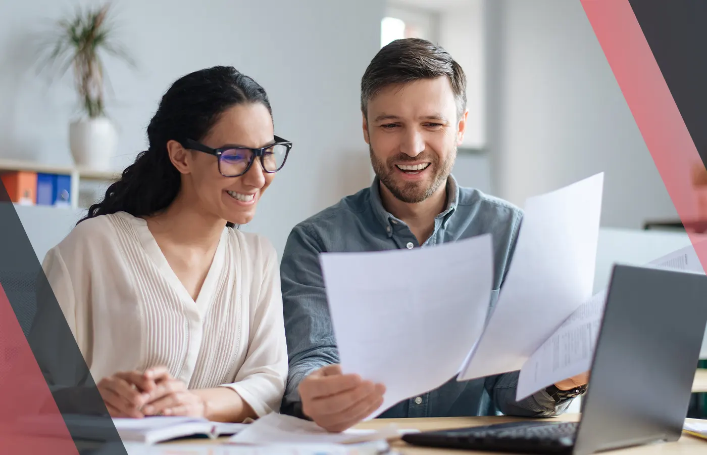 Two colleagues smiling while reviewing printed documents at a desk with a laptop in an office setting.