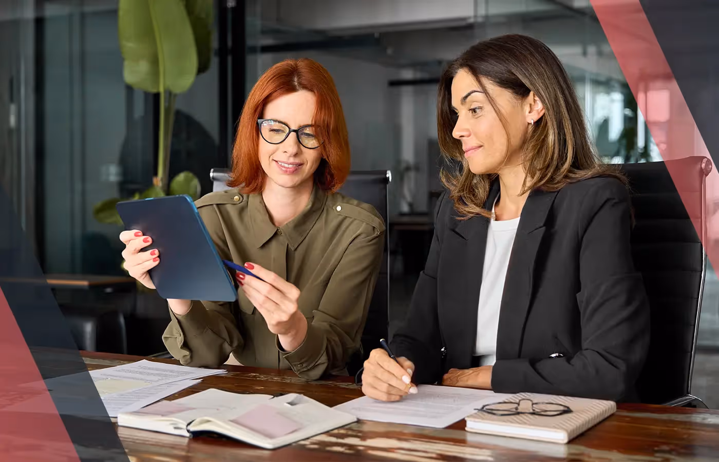 Two professionals in an office reviewing information on a tablet at a desk with papers and notebooks during a business discussion.